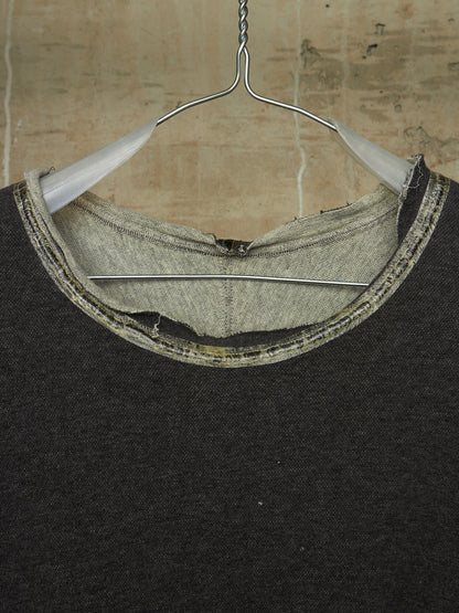 Close-up of a dark charcoal knit crewneck sweater hanging on a metal hanger against a textured beige wall. The neckline shows a raw, unfinished hem with a contrasting light gray inner lining and a stitched trim.