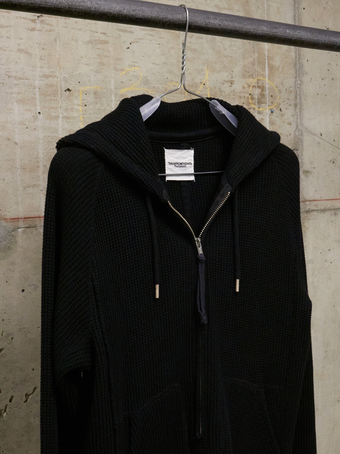 Black hooded coat hanging on a rack against a textured wall.