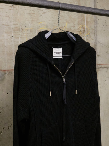 Black hooded coat hanging on a rack against a textured wall.