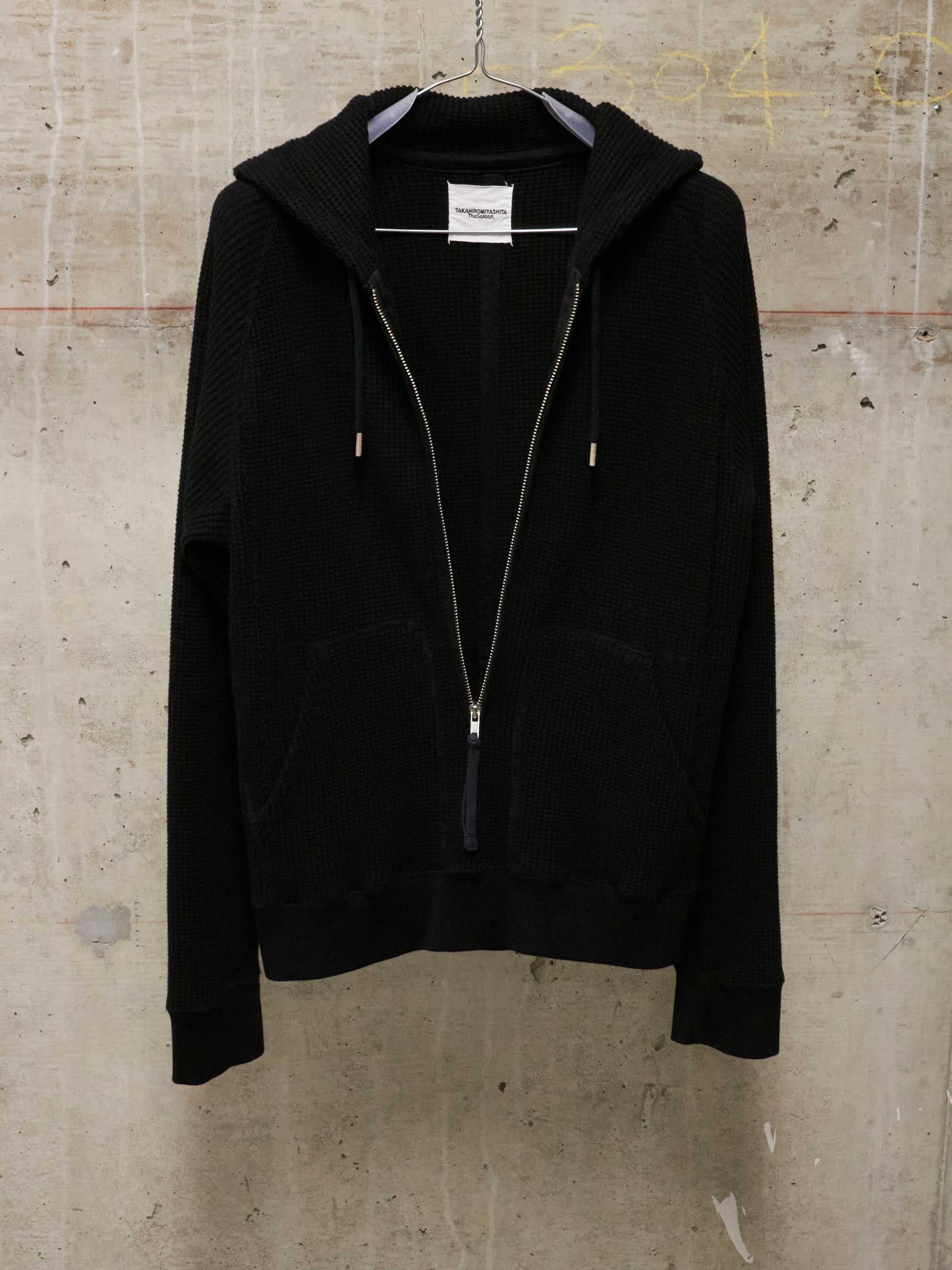 Black hoodie with a zipper on a hanger against a textured beige wall.