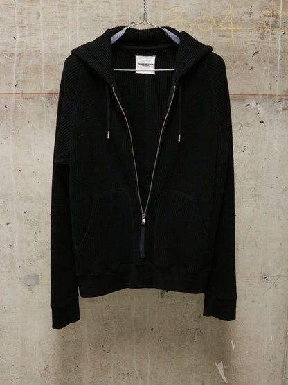 Black hoodie with a zipper on a hanger against a textured beige wall.