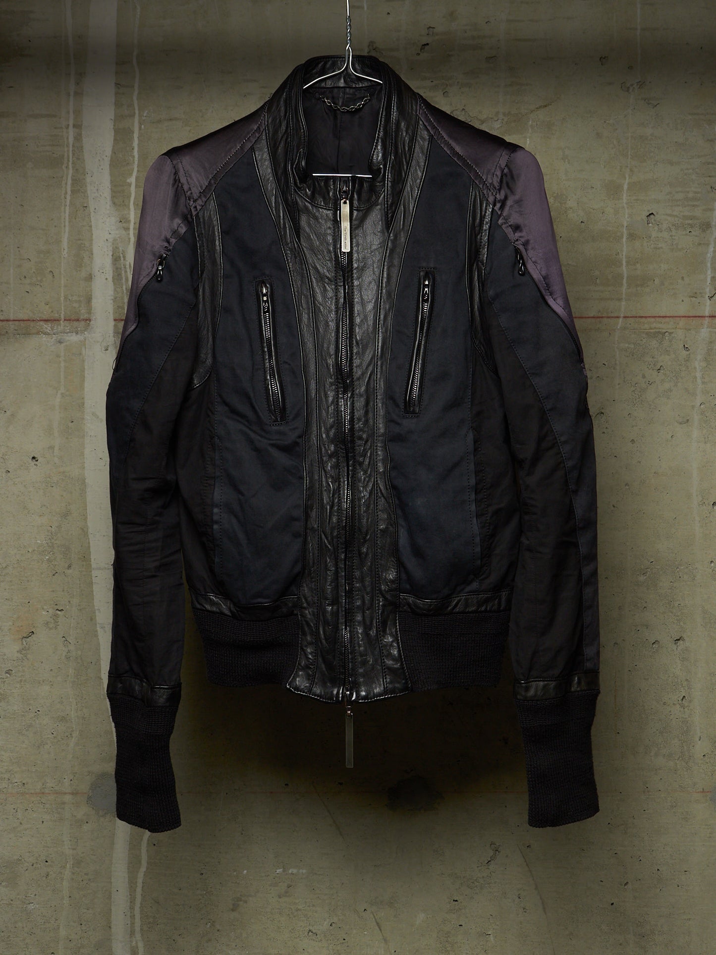 A dark bomber-style jacket with a glossy black leather front and matte black fabric body, featuring purple-toned shoulder panels, hanging against a textured concrete wall.
