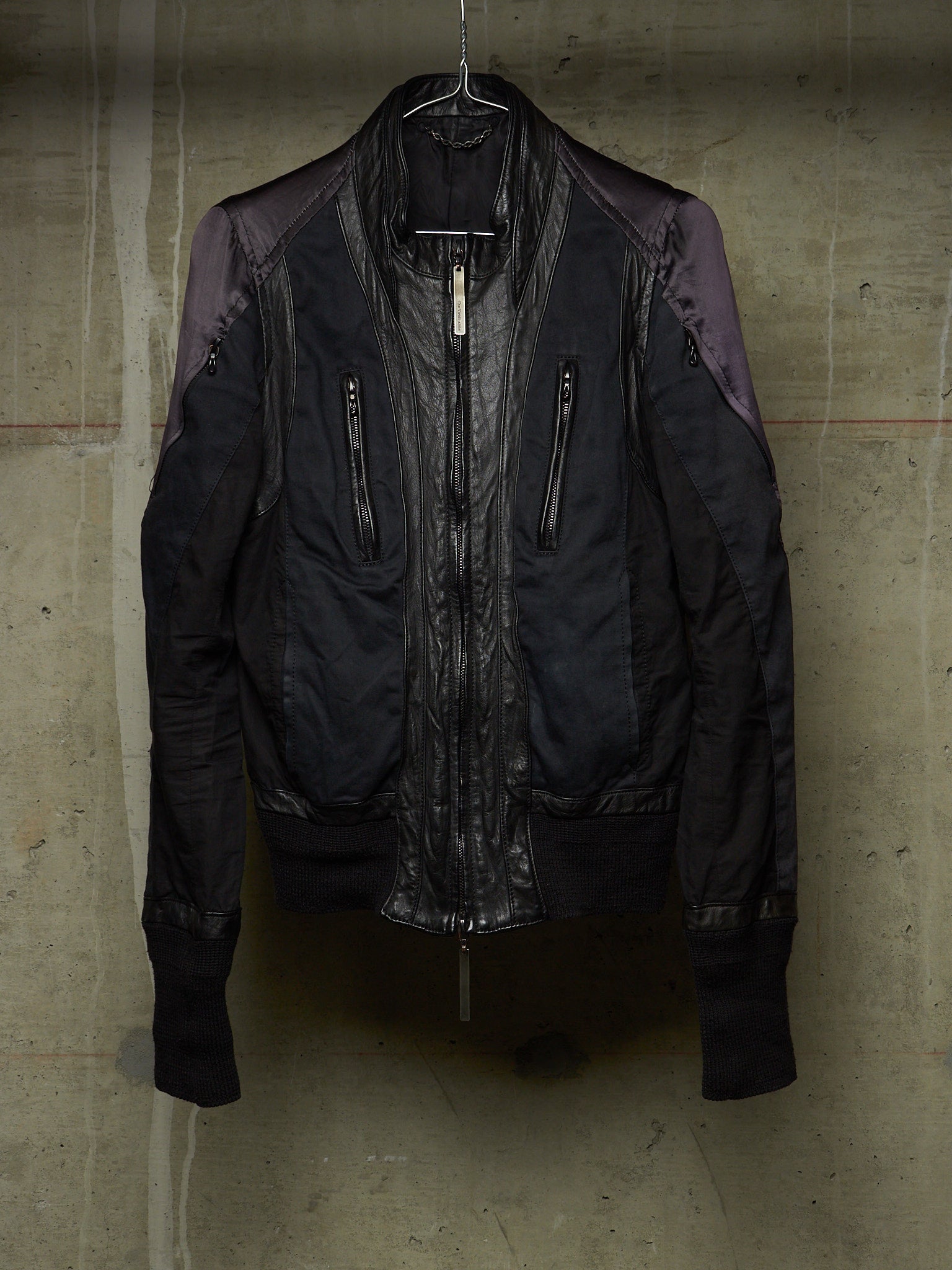 A dark bomber-style jacket with a glossy black leather front and matte black fabric body, featuring purple-toned shoulder panels, hanging against a textured concrete wall.