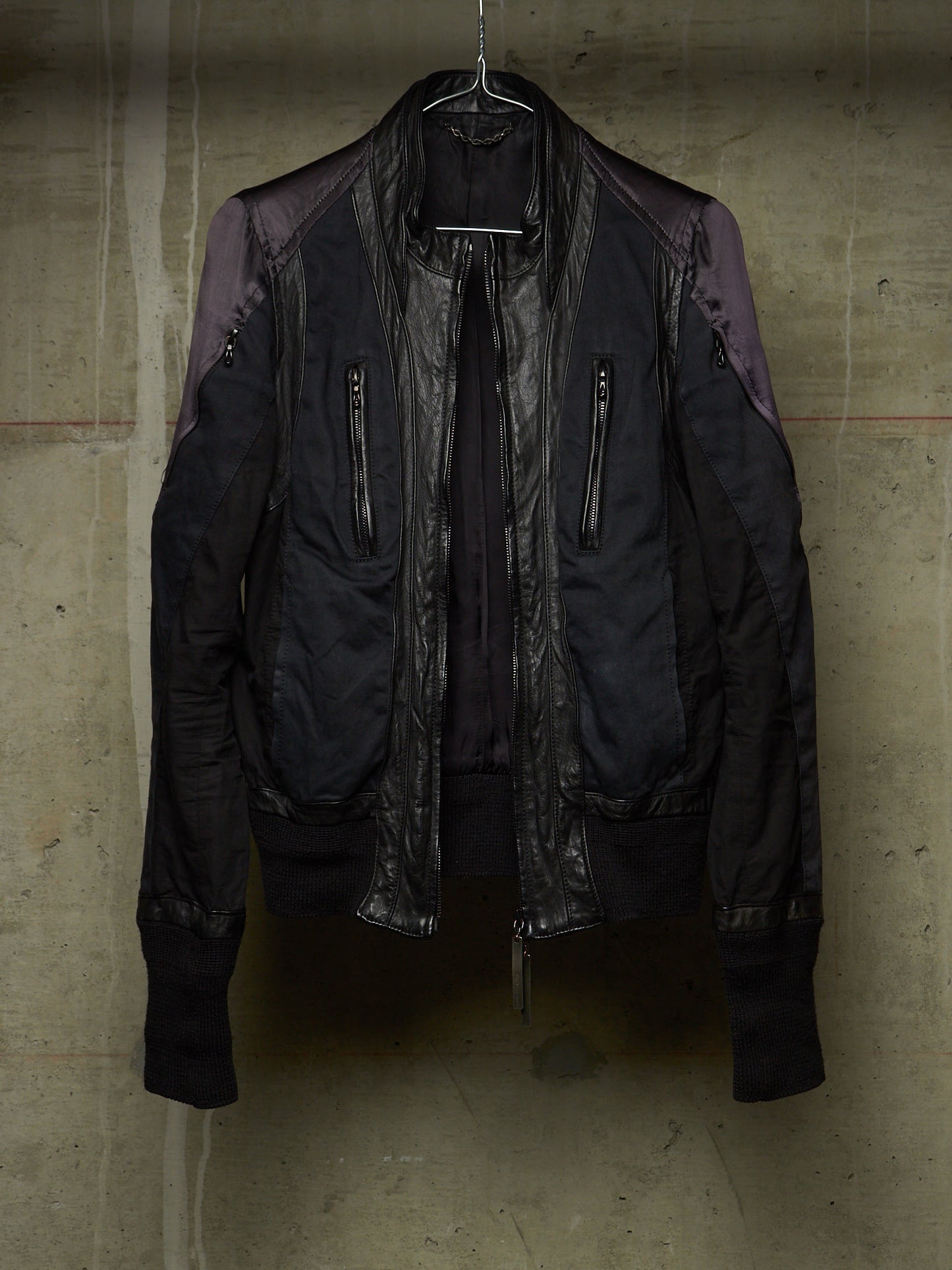A black bomber jacket with dark purple shoulder panels and a glossy black leather front panel, hanging on a wire hanger against a textured concrete wall.
