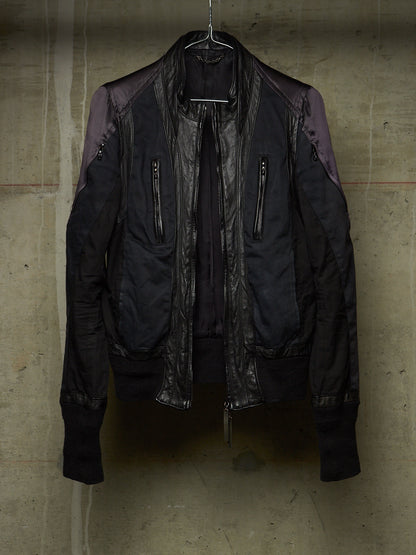 A black bomber jacket with dark purple shoulder panels and a glossy black leather front panel, hanging on a wire hanger against a textured concrete wall.