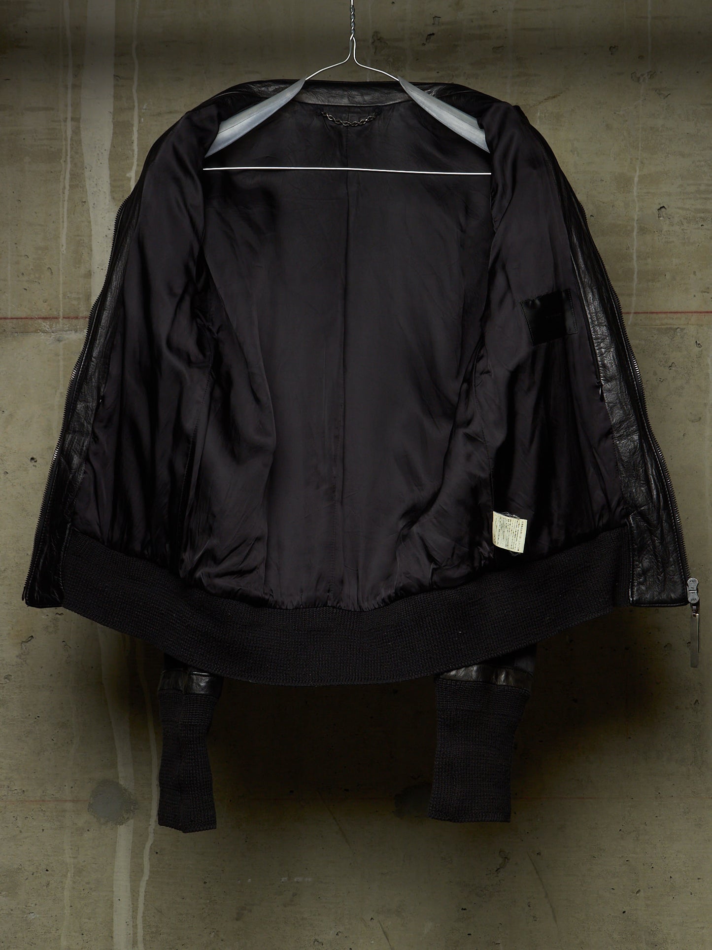 A black jacket with sheer mesh sleeves and a wide black collar, hanging on a metal hanger against a textured concrete wall.