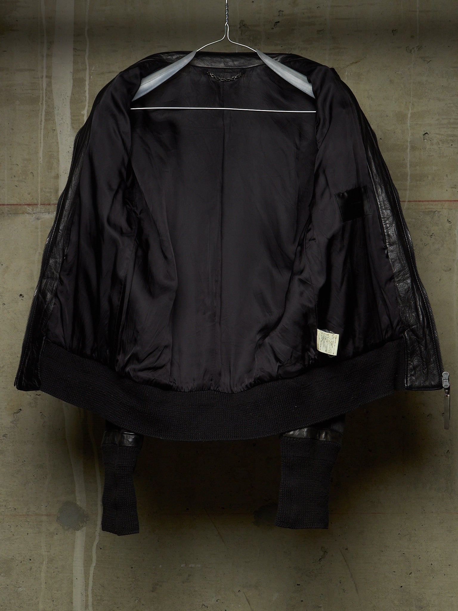 A black jacket with sheer mesh sleeves and a wide black collar, hanging on a metal hanger against a textured concrete wall.