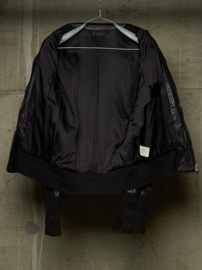 A black jacket with sheer mesh sleeves and a wide black collar, hanging on a metal hanger against a textured concrete wall.