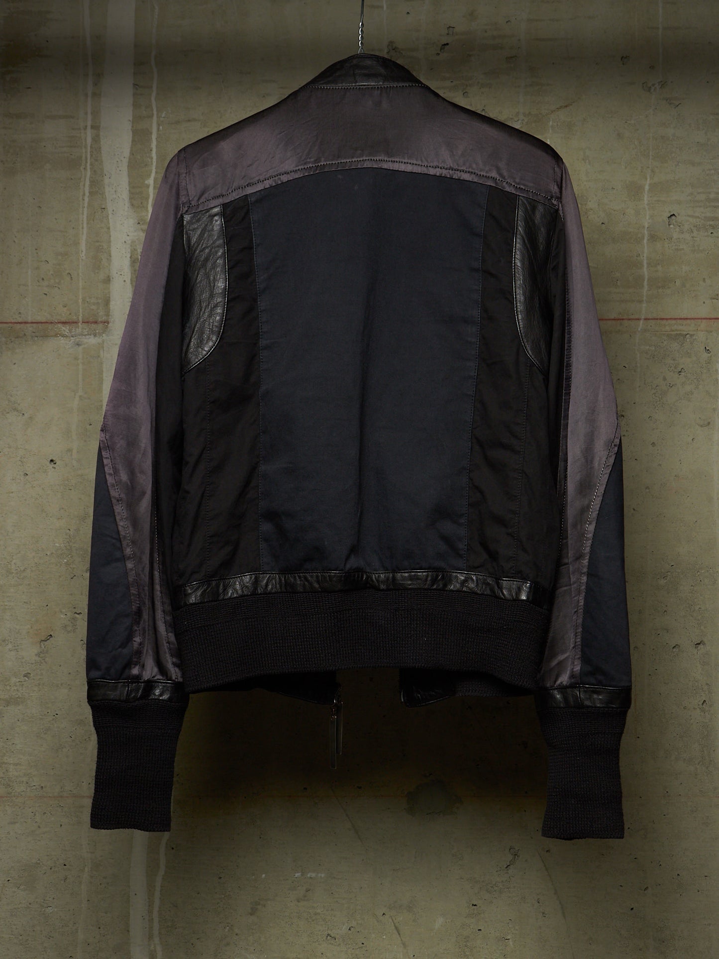 A dark bomber jacket with contrasting gray-purple sleeves and back panels, hanging against a textured concrete wall.