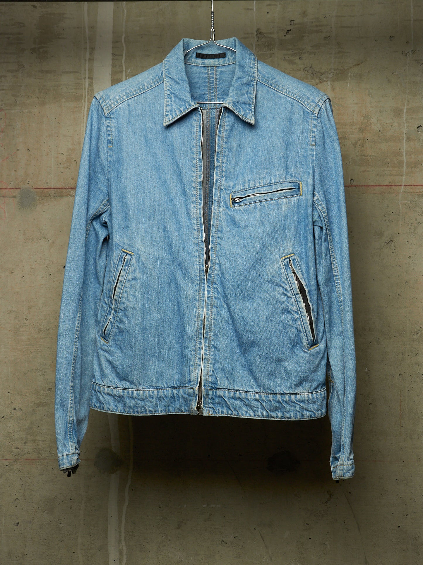 A light blue denim jacket hanging against a textured concrete wall. The jacket features a pointed collar, a front zipper closure, a zippered chest pocket, and two zippered hand pockets.
