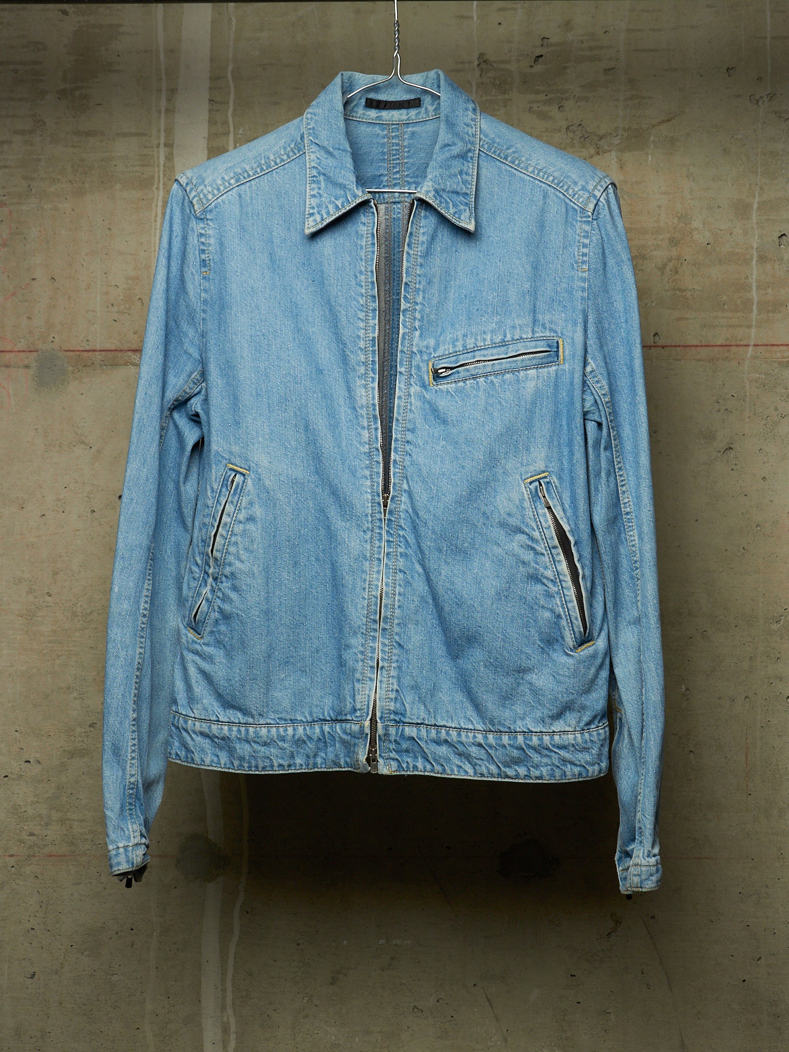 A light blue denim jacket hanging against a textured concrete wall. The jacket features a pointed collar, a front zipper closure, a zippered chest pocket, and two zippered hand pockets.