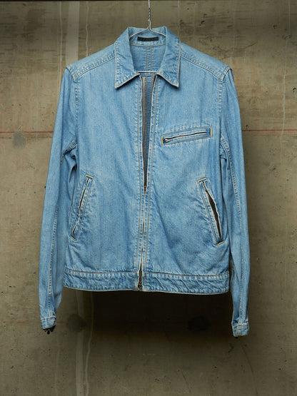 A light blue denim jacket hanging against a textured concrete wall. The jacket features a pointed collar, a front zipper closure, a zippered chest pocket, and two zippered hand pockets.