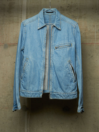 A light blue denim jacket hanging against a textured concrete wall, featuring a front zipper closure, zippered chest pocket, and zippered side pockets.