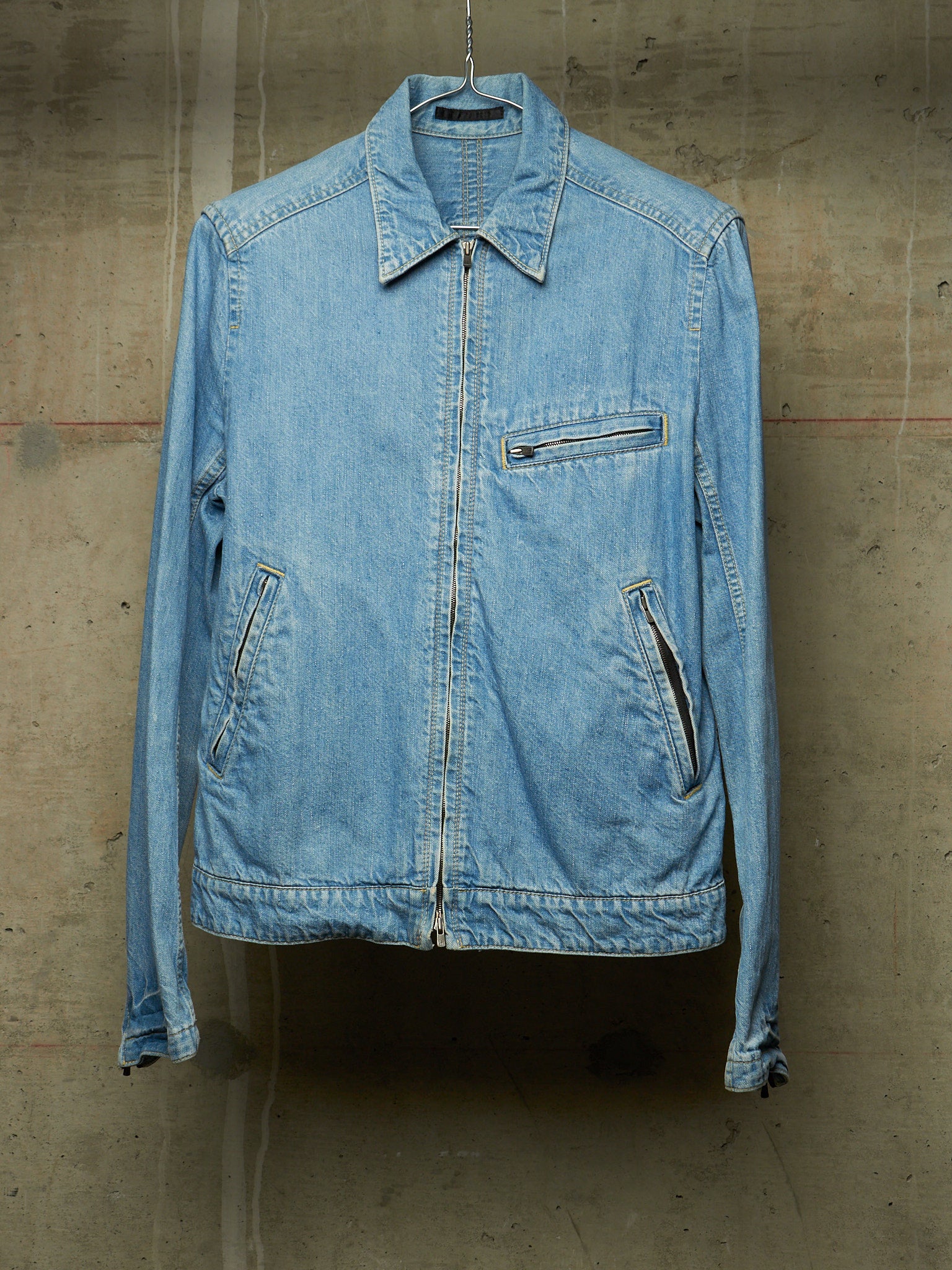 A light blue denim jacket hanging on a metal hanger against a textured concrete wall.