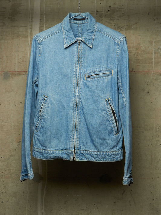 A light blue denim jacket hanging on a metal hanger against a textured concrete wall.