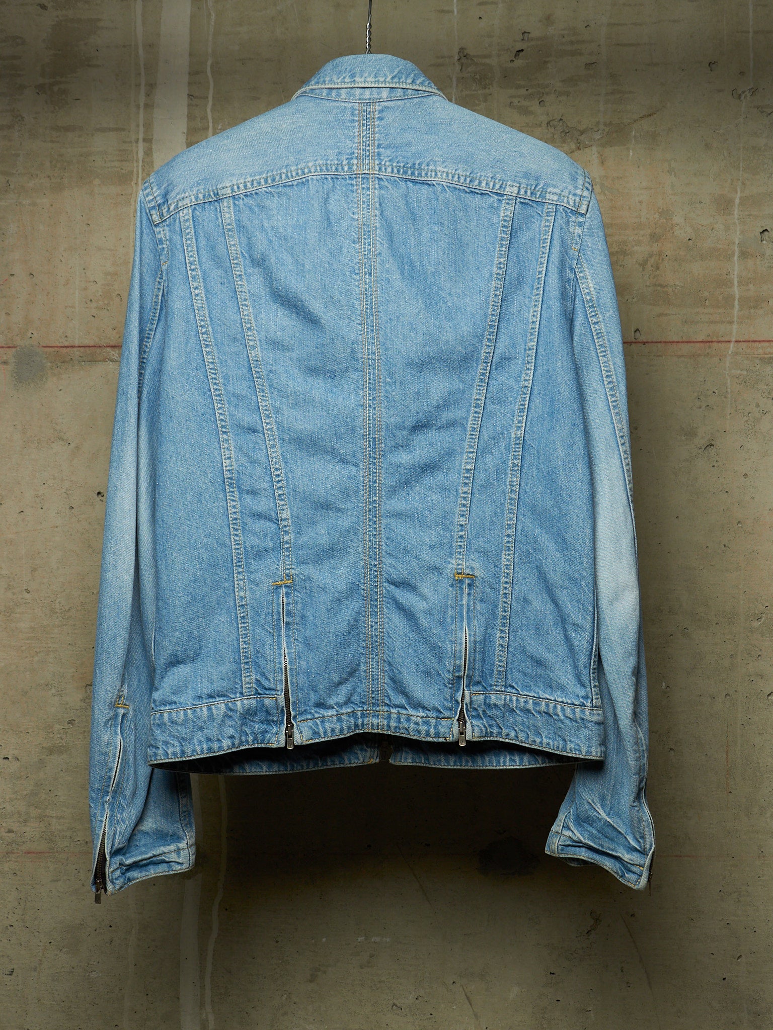 A light blue denim jacket shown from the back, hanging against a textured concrete wall.