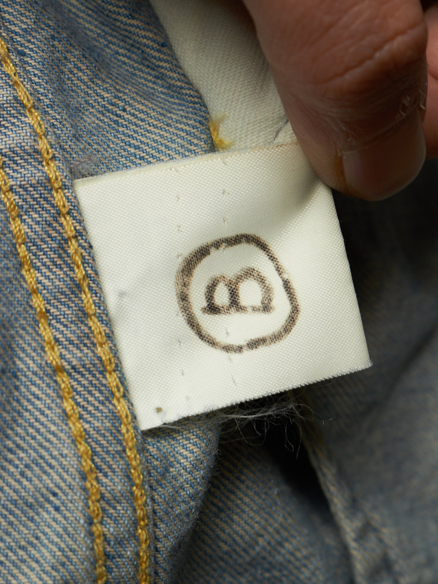 Close-up of a white fabric sewn-in size tag attached to blue denim clothing, with a finger holding the tag in place. The tag shows a dark brown circle-with-two-b symbol, and the denim has yellow contrast stitching.
