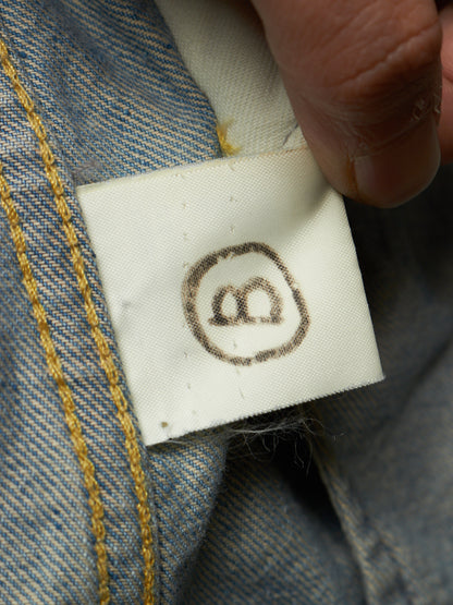 Close-up of a white fabric sewn-in size tag attached to blue denim clothing, with a finger holding the tag in place. The tag shows a dark brown circle-with-two-b symbol, and the denim has yellow contrast stitching.