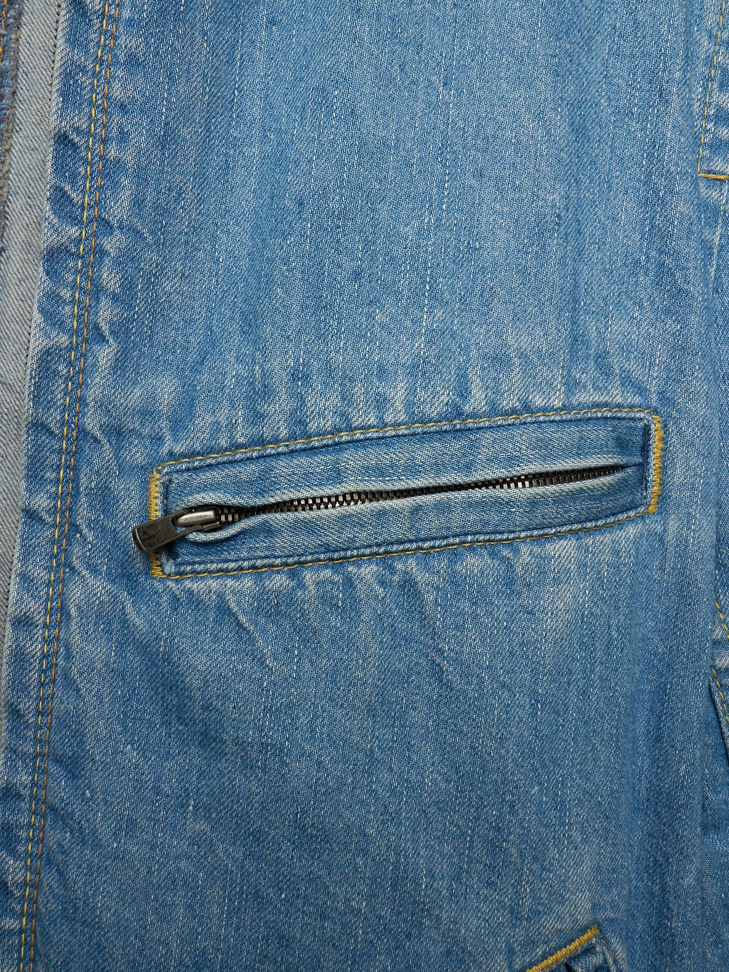Close-up of a light blue denim jacket featuring a zippered pocket with a black zipper pull and contrast yellow stitching.