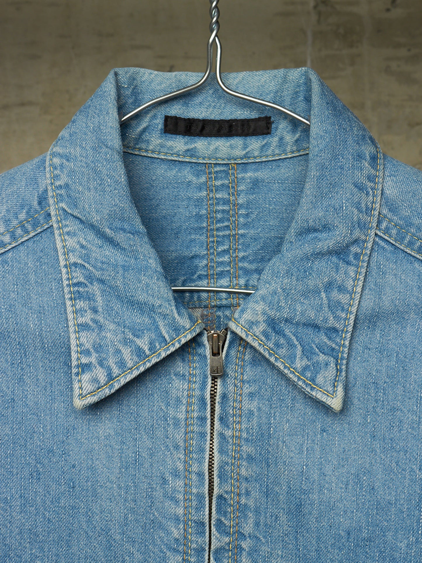 A close-up of a light blue denim jacket hanging on a metal hanger against a textured gray wall. The jacket features a pointed collar, a front zipper, and detailed contrast stitching.