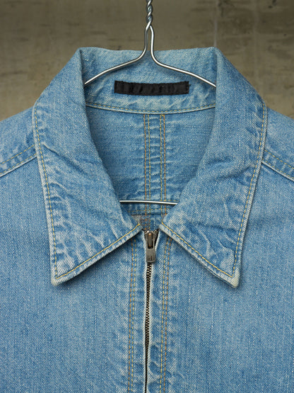 A close-up of a light blue denim jacket hanging on a metal hanger against a textured gray wall. The jacket features a pointed collar, a front zipper, and detailed contrast stitching.