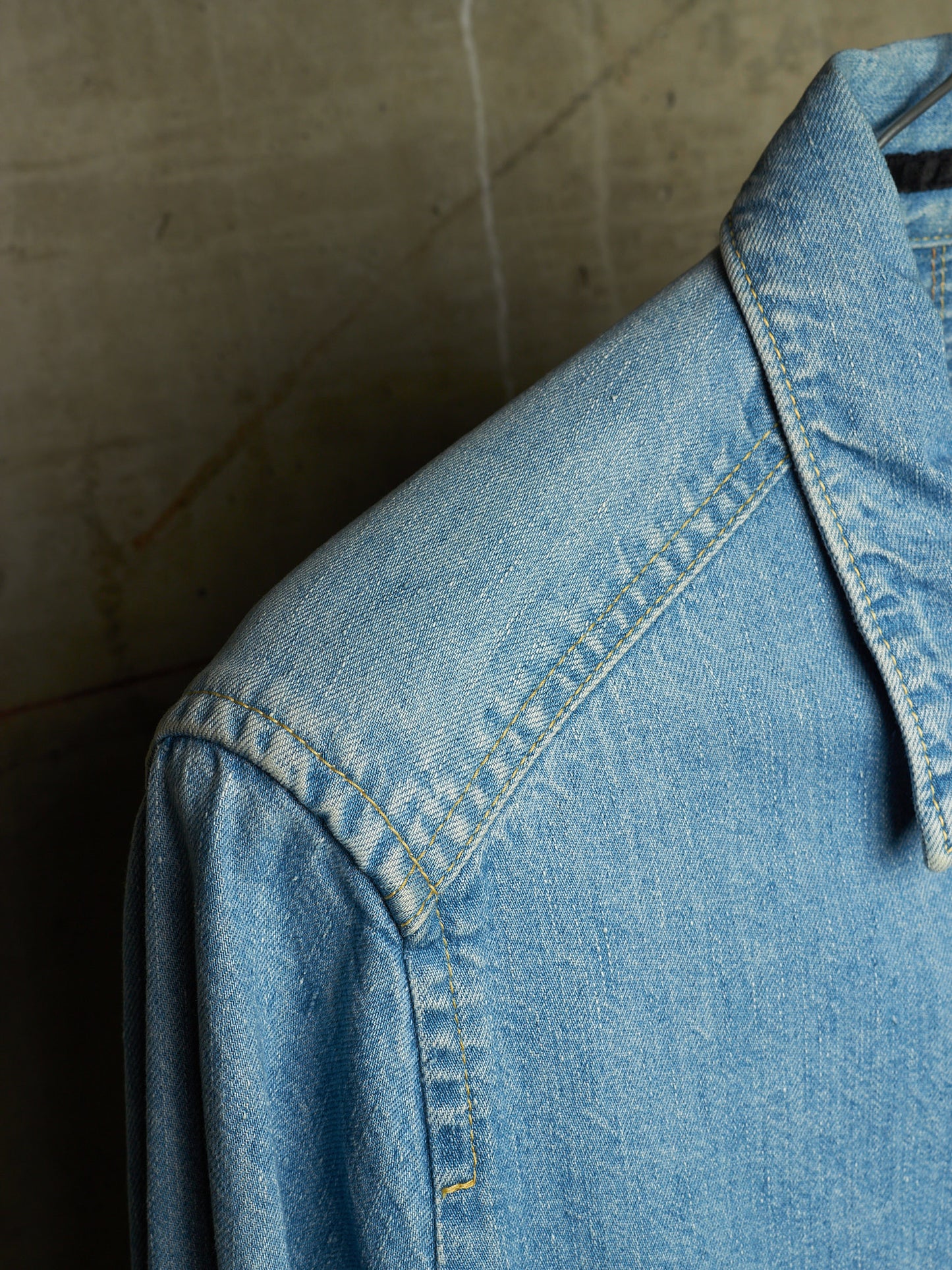 Close-up of a light blue denim shirt jacket showing the collar, shoulder, and detailed stitching against a textured concrete background.