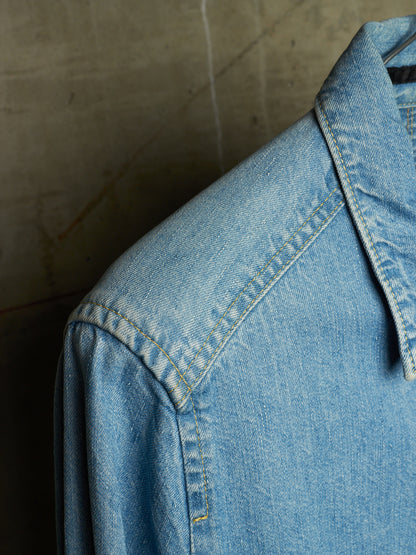 Close-up of a light blue denim shirt jacket showing the collar, shoulder, and detailed stitching against a textured concrete background.