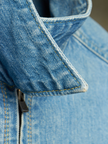 Close-up detail of a light blue denim jacket showing the collar, stitching, and a zipper pull.