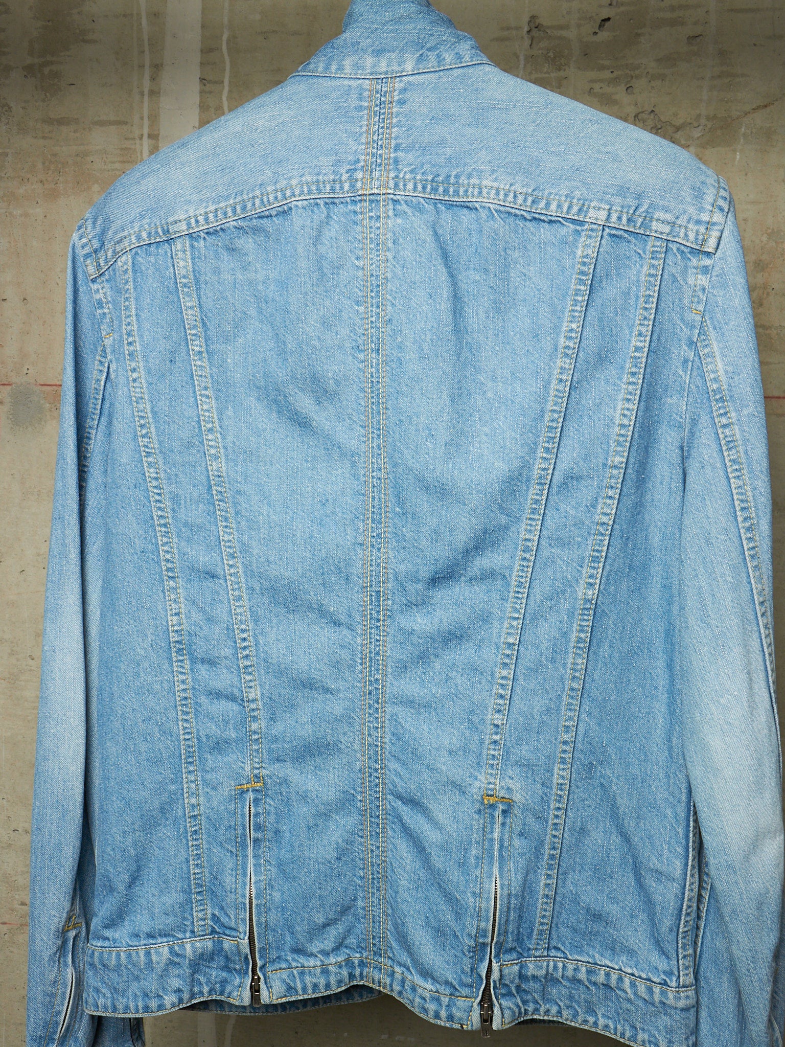 A light blue denim jacket shown from the back, featuring a central seam, zippered back pockets, and a collar, photographed against a textured concrete wall.