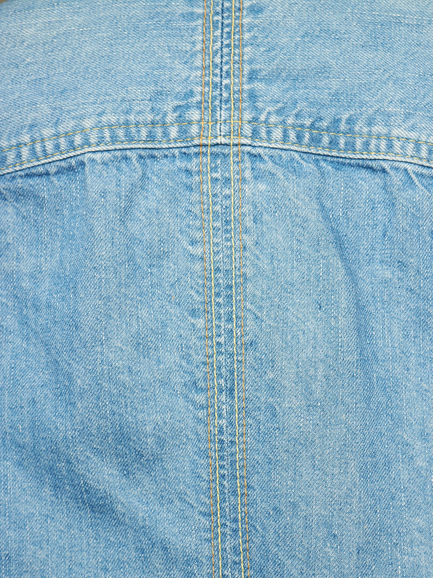 A close-up of light blue denim fabric showing the texture of the weave and a central double-stitched seam.