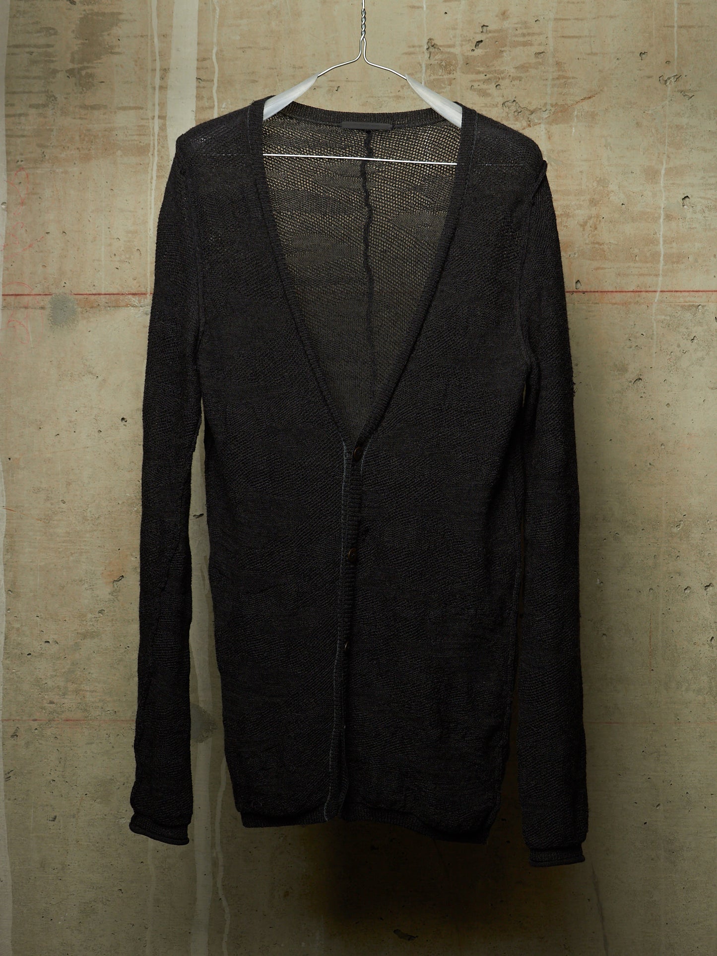 A black open-front cardigan sweater with a deep V-neck and long sleeves, hanging on a white plastic hanger against a textured concrete wall.