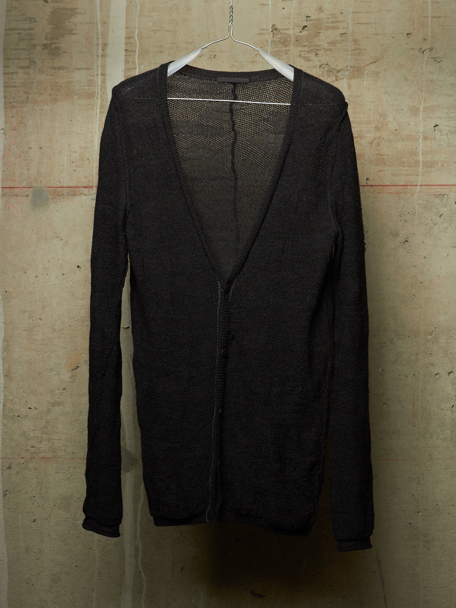 A black open-front cardigan sweater with a deep V-neck and long sleeves, hanging on a white plastic hanger against a textured concrete wall.
