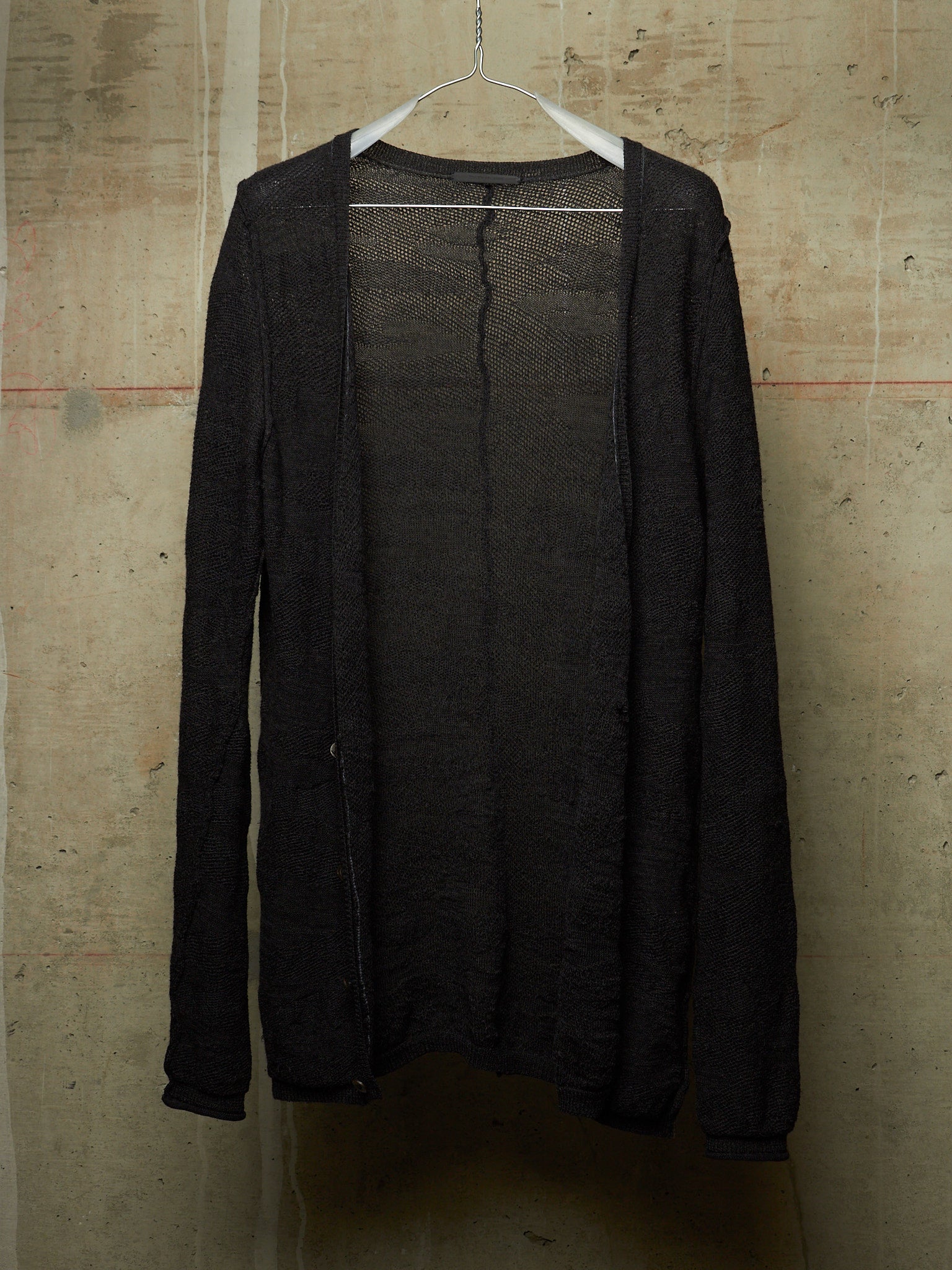 A black open-front knit cardigan sweater hanging on a white plastic hanger against a textured concrete wall.