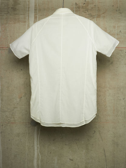 A white short-sleeve button-up shirt shown from the back, hanging against a textured concrete wall.