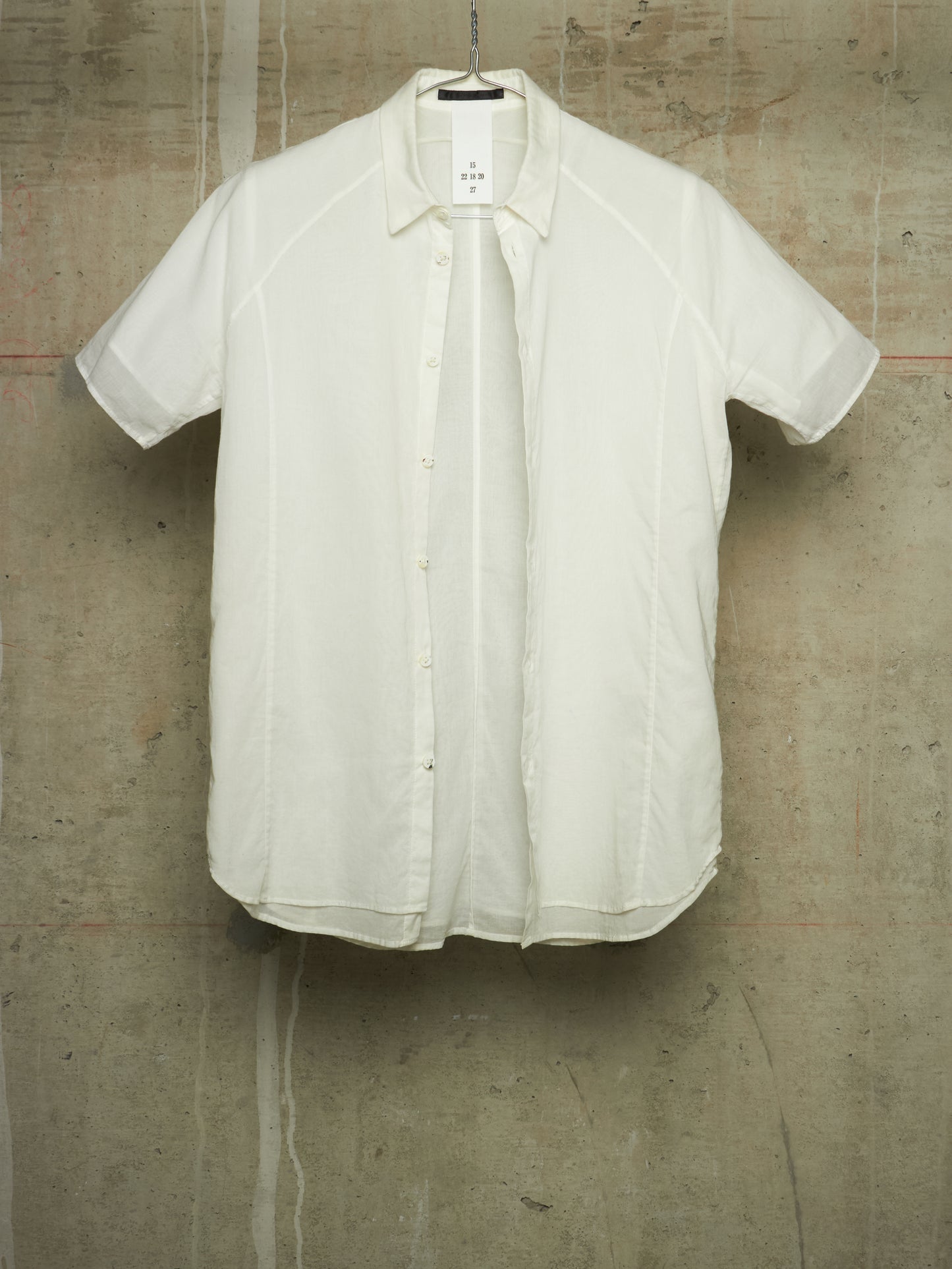 A short-sleeve button-up shirt in off-white/cream fabric, hanging against a textured concrete wall.