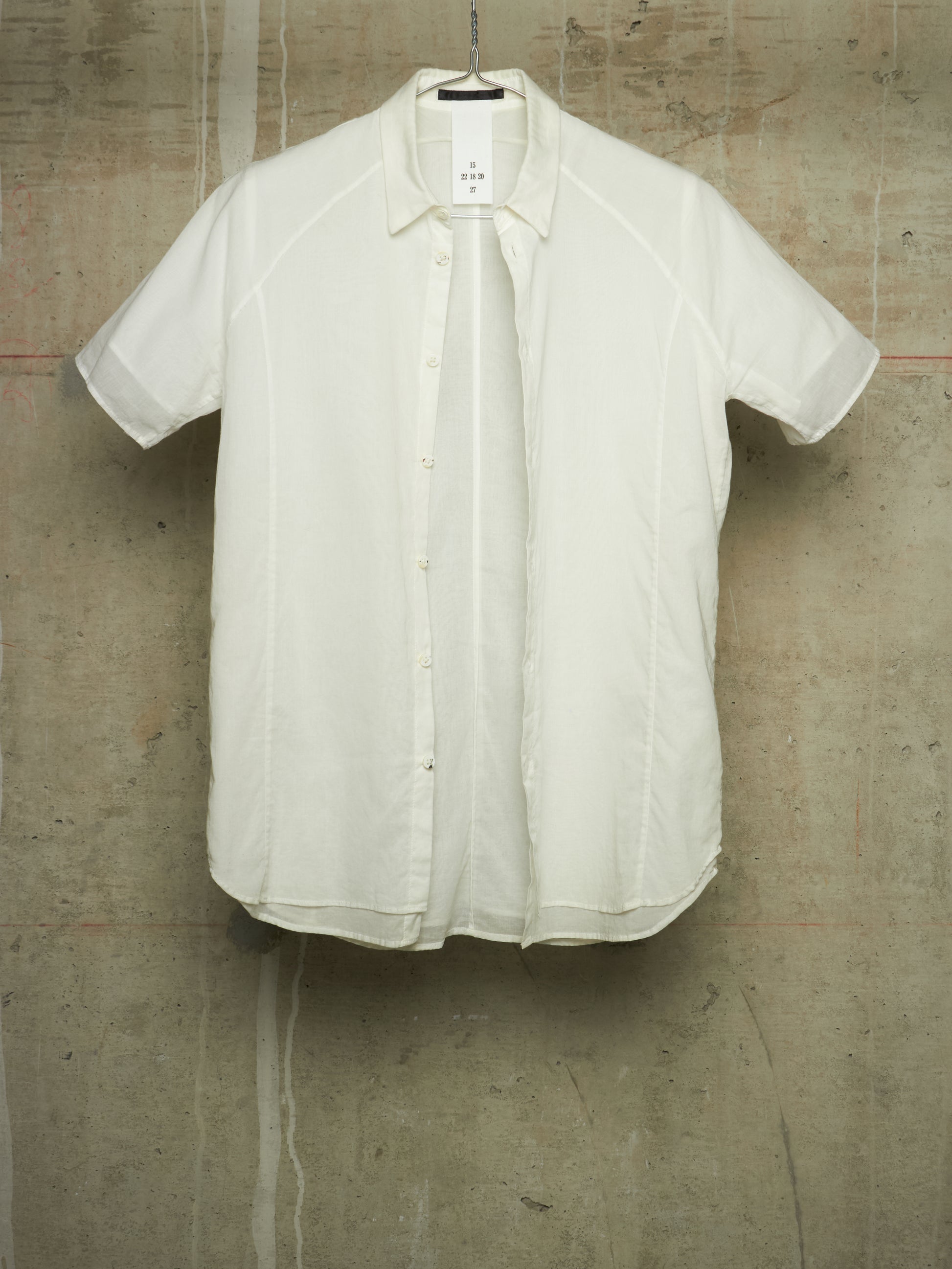 A short-sleeve button-up shirt in off-white/cream fabric, hanging against a textured concrete wall.