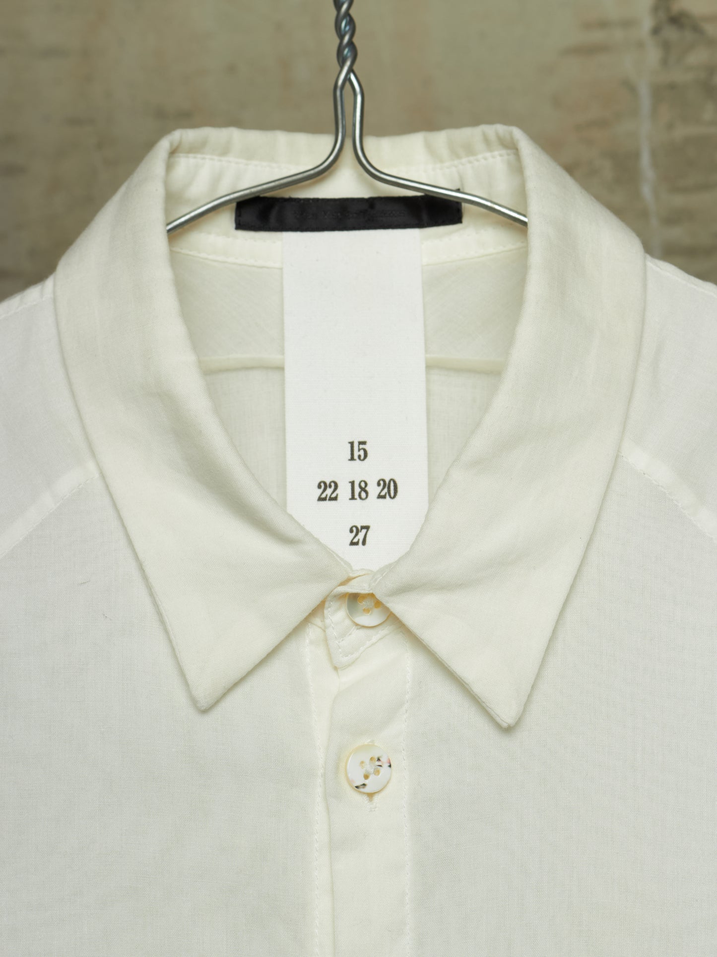 Close-up of a cream-colored knit collared shirt hanging on a metal hanger, with a white size tag sewn into the collar showing the numbers 15, 22 18 20, and 27.