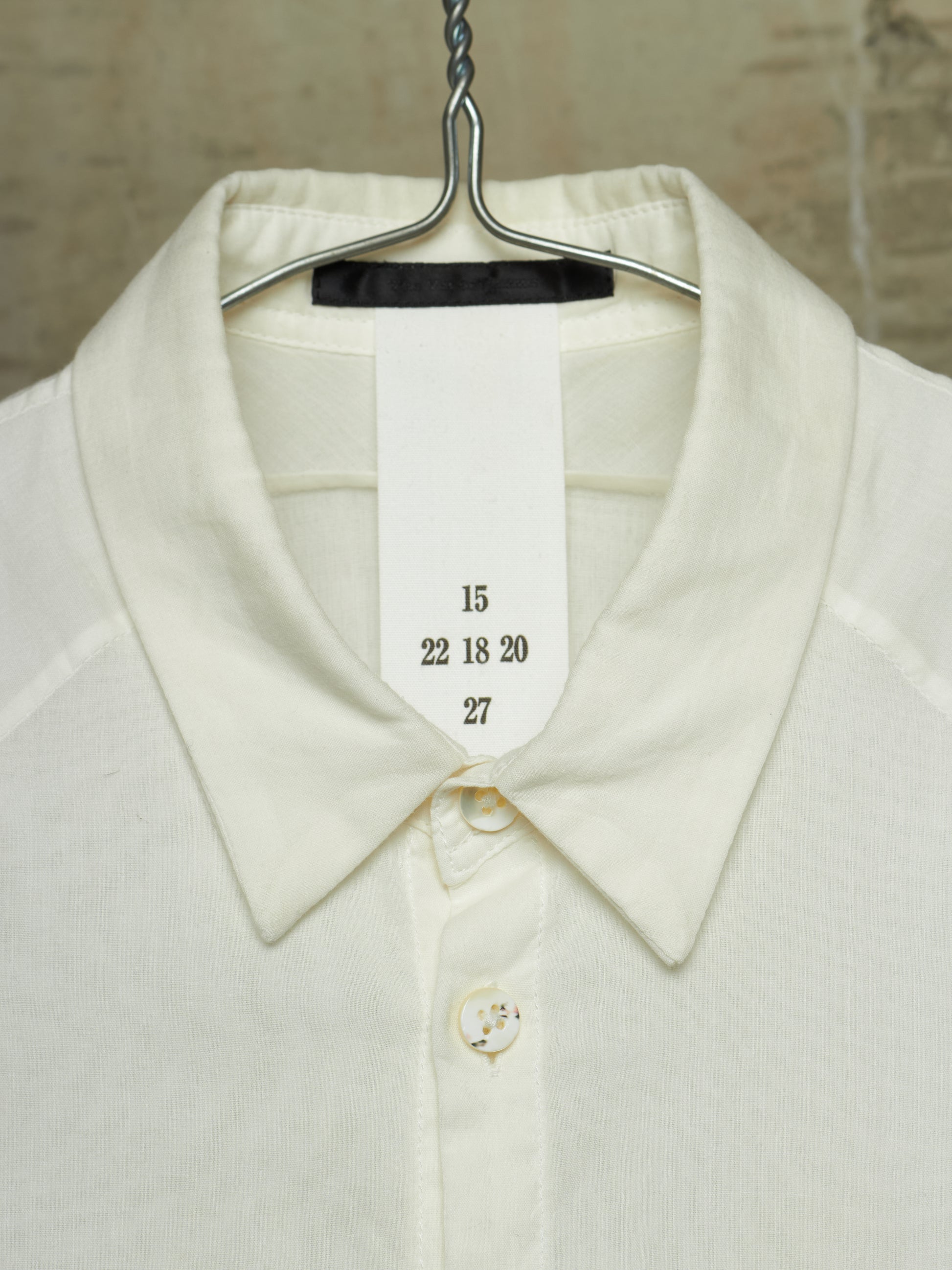 Close-up of a cream-colored knit collared shirt hanging on a metal hanger, with a white size tag sewn into the collar showing the numbers 15, 22 18 20, and 27.