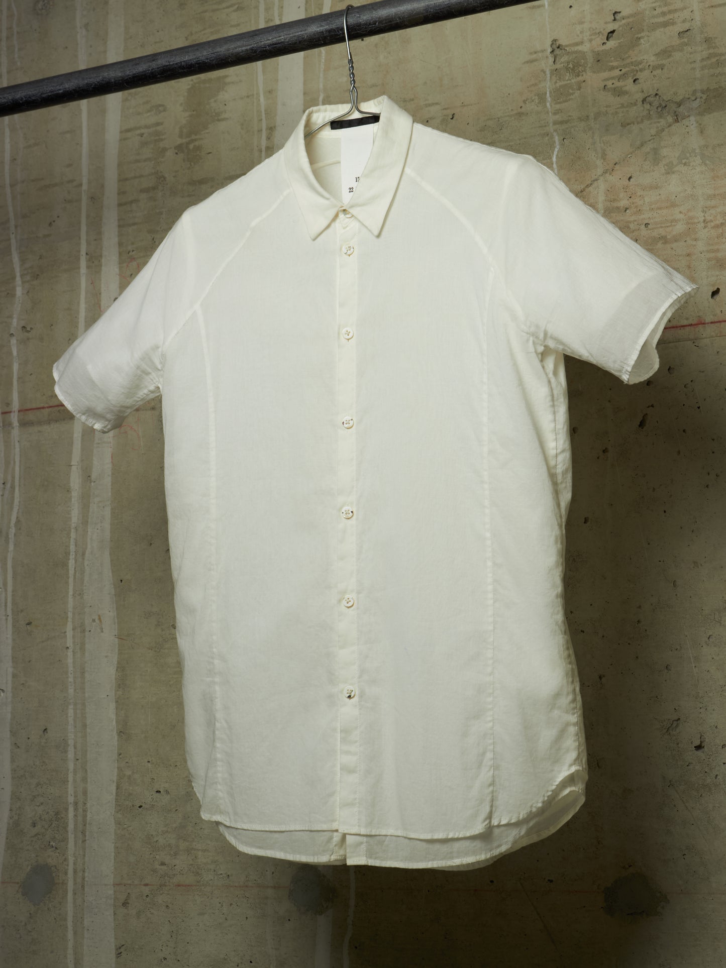 An off-white short-sleeve button-up shirt with a relaxed, oversized fit, hanging on a black hanger from a metal rod against a textured concrete wall.