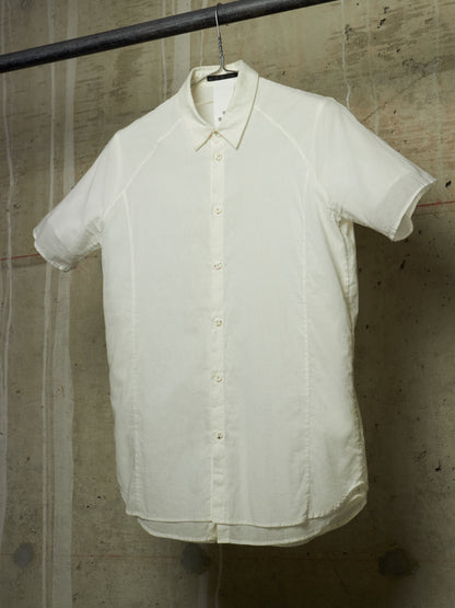 An off-white short-sleeve button-up shirt with a relaxed, oversized fit, hanging on a black hanger from a metal rod against a textured concrete wall.