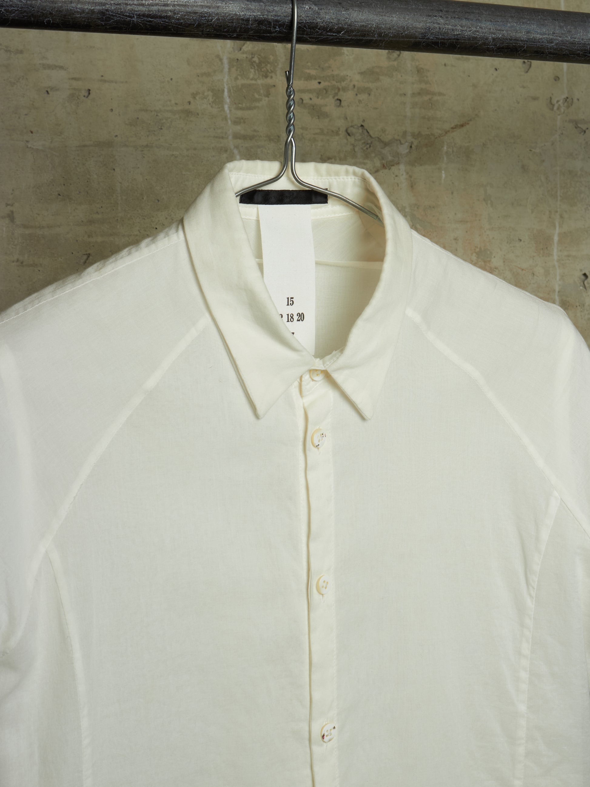 An off-white long-sleeve button-up shirt with a pointed collar, hanging on a black metal hanger from a dark industrial rod against a textured concrete wall.