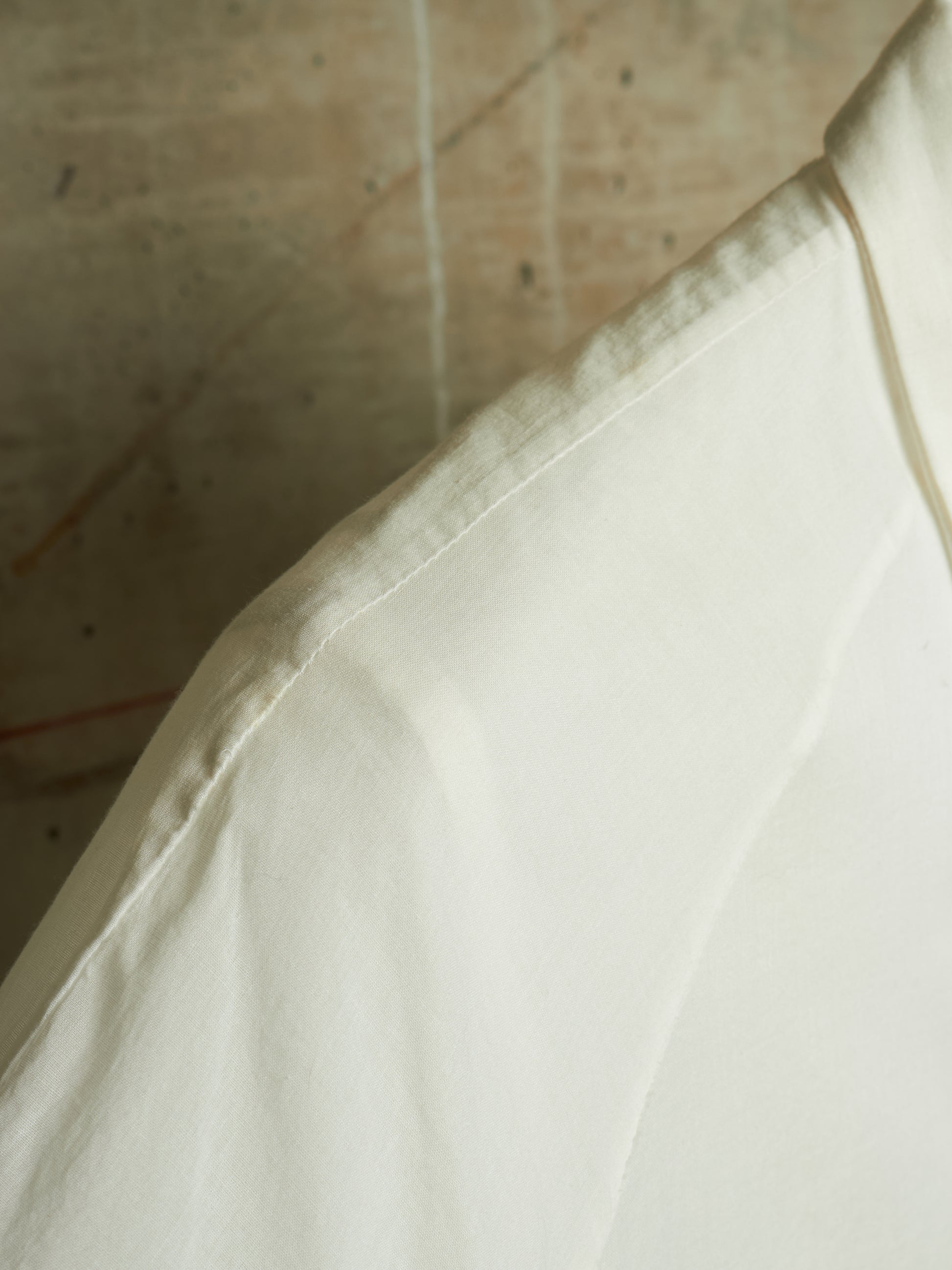 A close-up detail shot of a white or off-white fabric garment, showing the shoulder seam and collar area against a textured, warm-toned background.