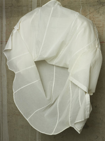 An off-white, semi-sheer linen garment with vertical stitched panel lines, draped to show its back design against a textured concrete wall.