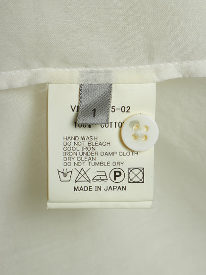 Close-up of a garment's care label attached to a white fabric hem, showing text including '100% COTTON', care symbols, and 'MADE IN JAPAN' with a white button nearby.