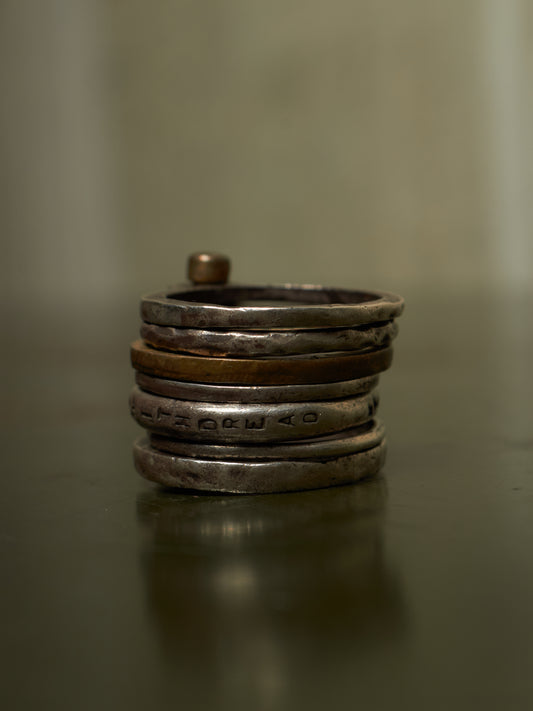 A stack of handmade silver rings, including one engraved with Hebrew script and one with a small copper-colored accent, displayed on a dark reflective surface.