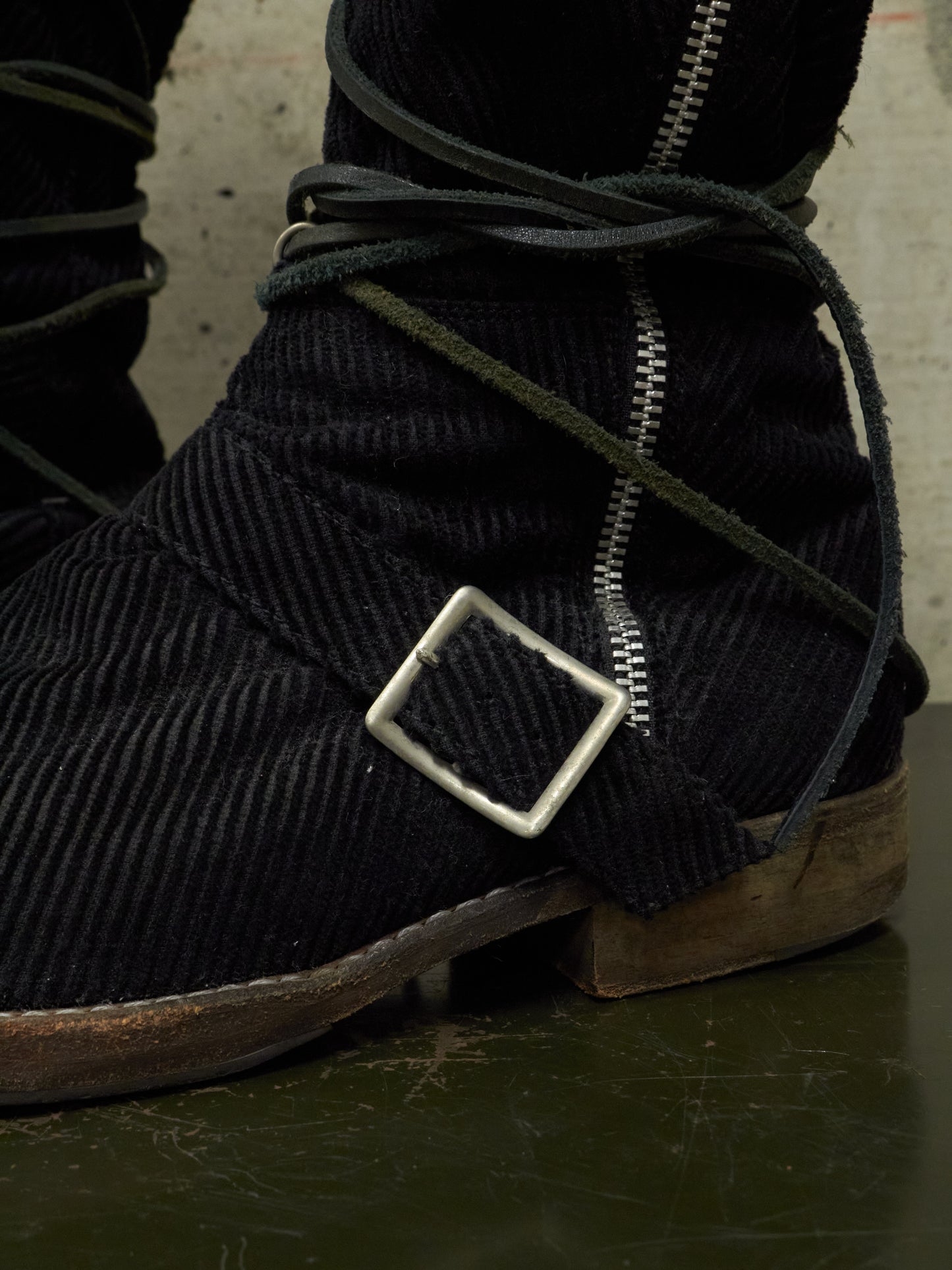 Close-up of dark corduroy boots with a metal buckle and zipper on a textured surface.