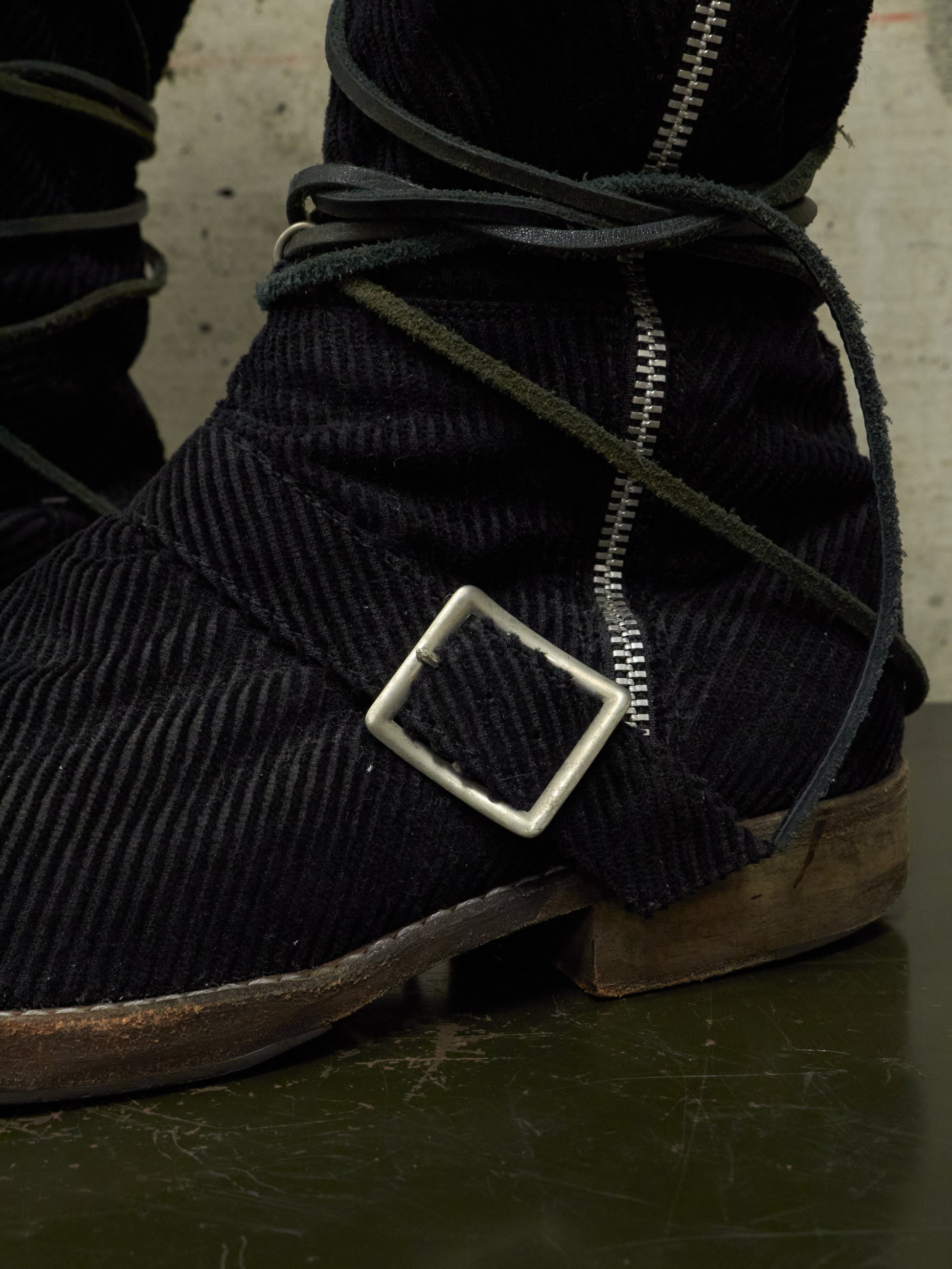 Close-up of dark corduroy boots with a metal buckle and zipper on a textured surface.