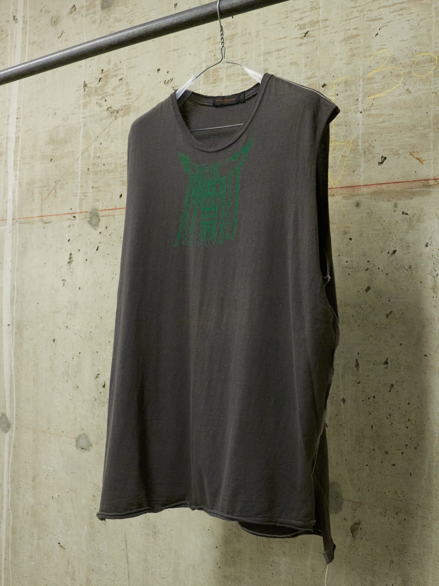 Gray sleeveless shirt with green logo on a hanger against a concrete wall.