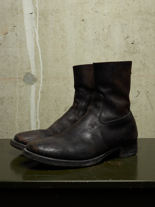 A pair of dark brown distressed leather ankle boots with a rugged, worn finish, displayed on a dark green surface against a textured concrete wall.