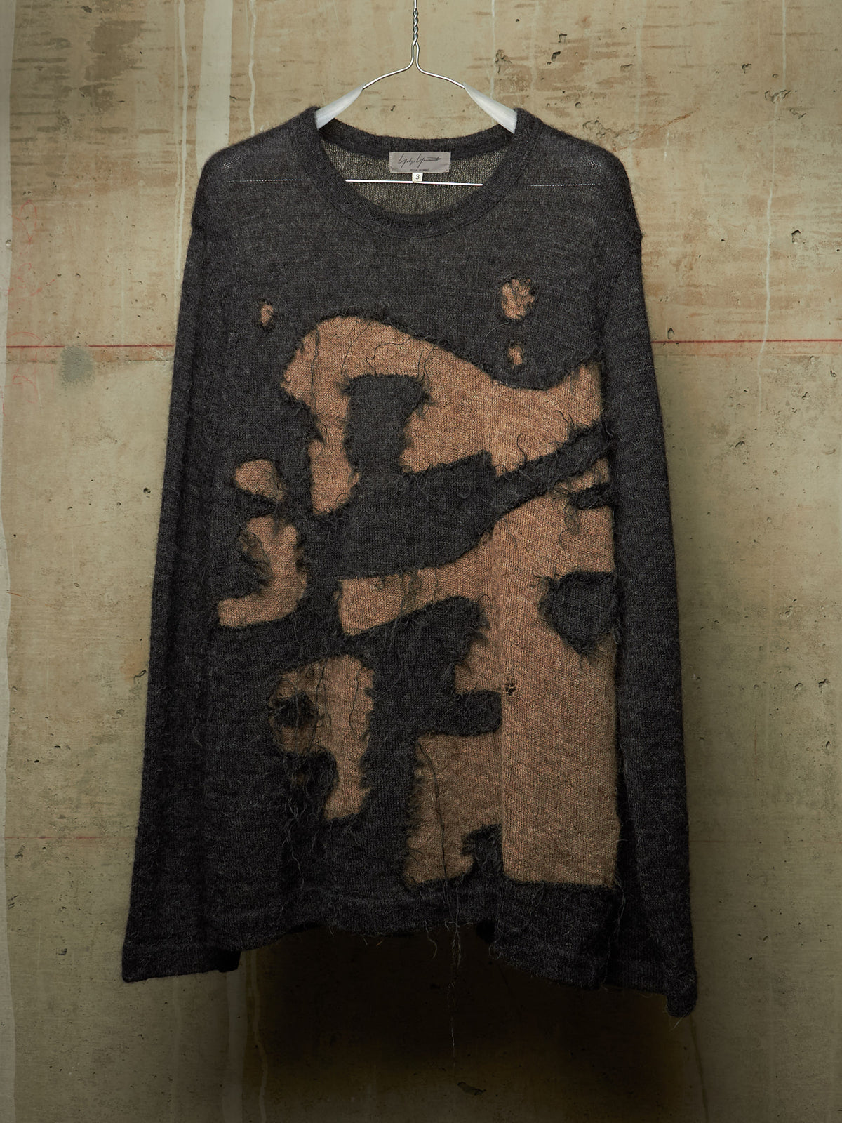 A dark charcoal gray long-sleeve knit sweater with a large abstract tan and brown distressed graphic on the front, hanging on a white wire hanger against a textured concrete wall.